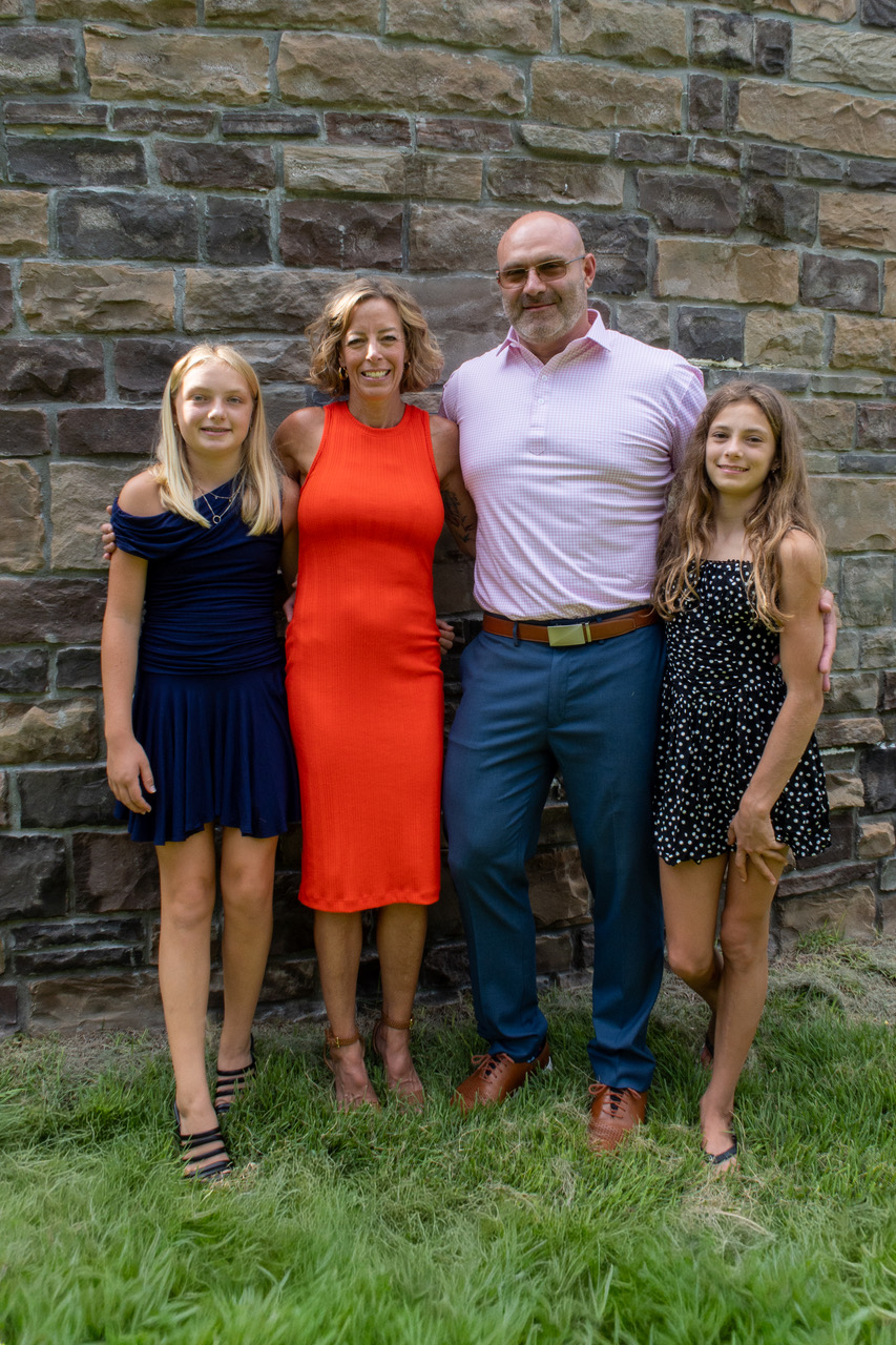 Mitchel and Jessica Krause standing outdoors with their two daughters in front of a stone wall.
